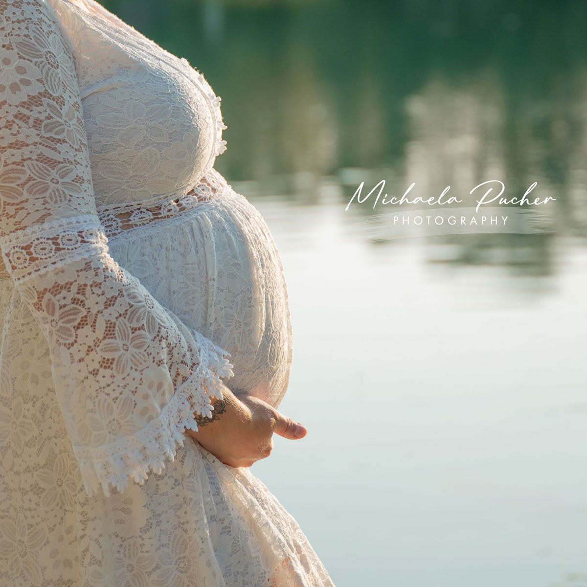Babybauch in einem weißen, bestickten Spitzenkleid im warmen Abendlicht am Wasser.